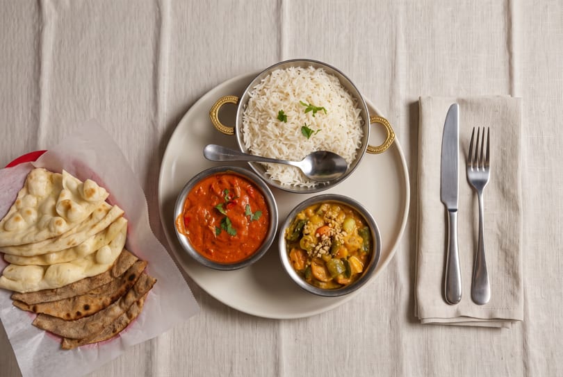 Overhead view of an Indian meal with rice, two curries, naan, and roti on a white tablecloth.