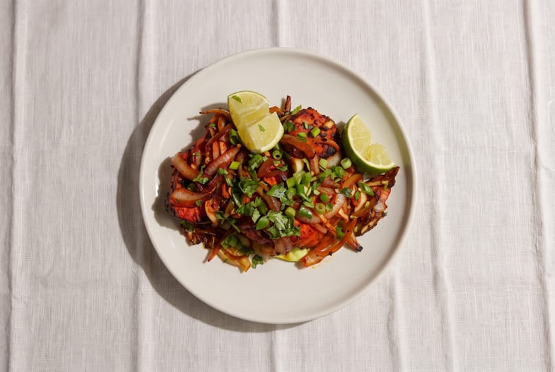Tandoori chicken with sautéed onions, cilantro, and lime wedges on a white plate.